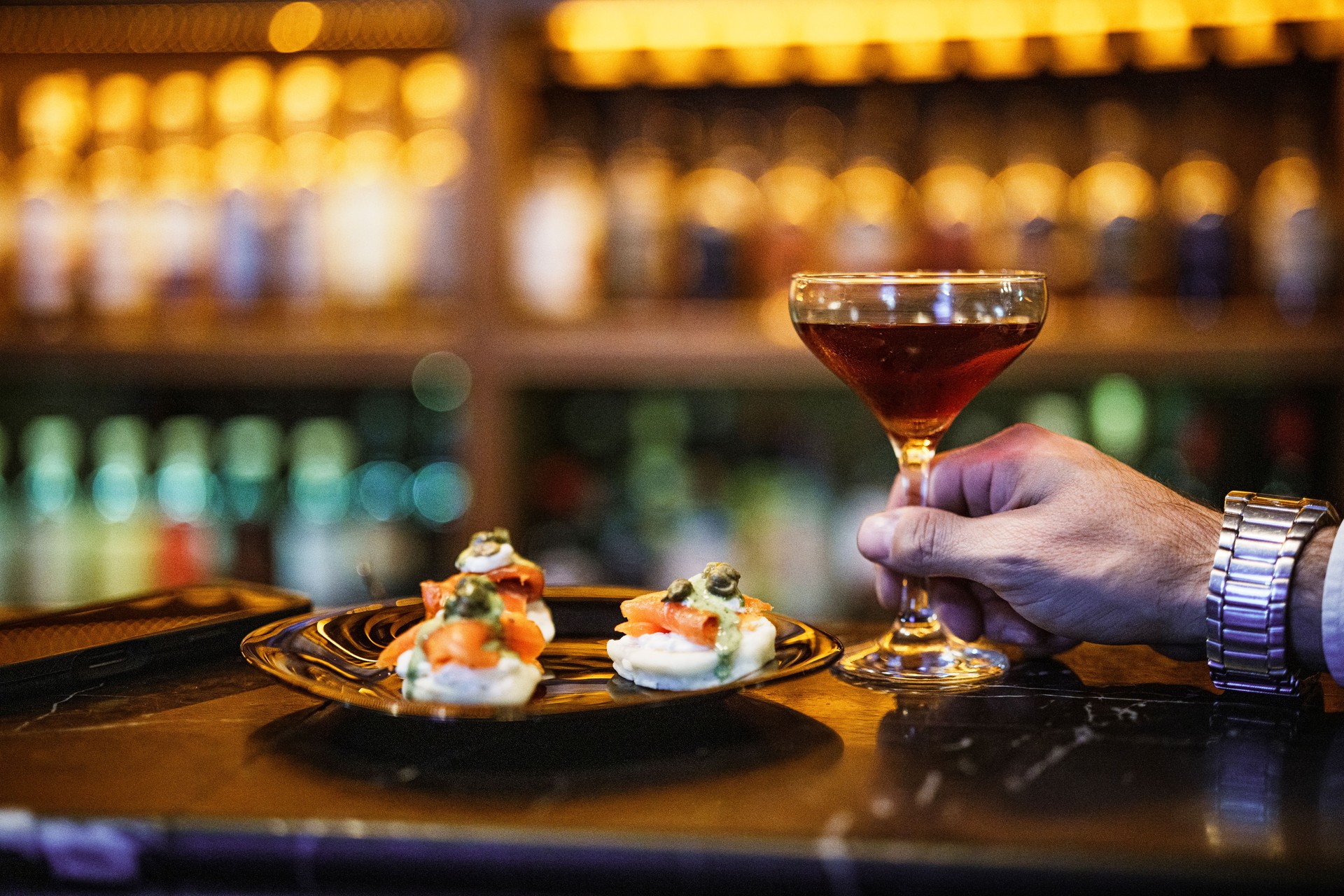 Close-up shot man's hand holding a Manhattan cocktail next to salmon tapas at luxury bar.