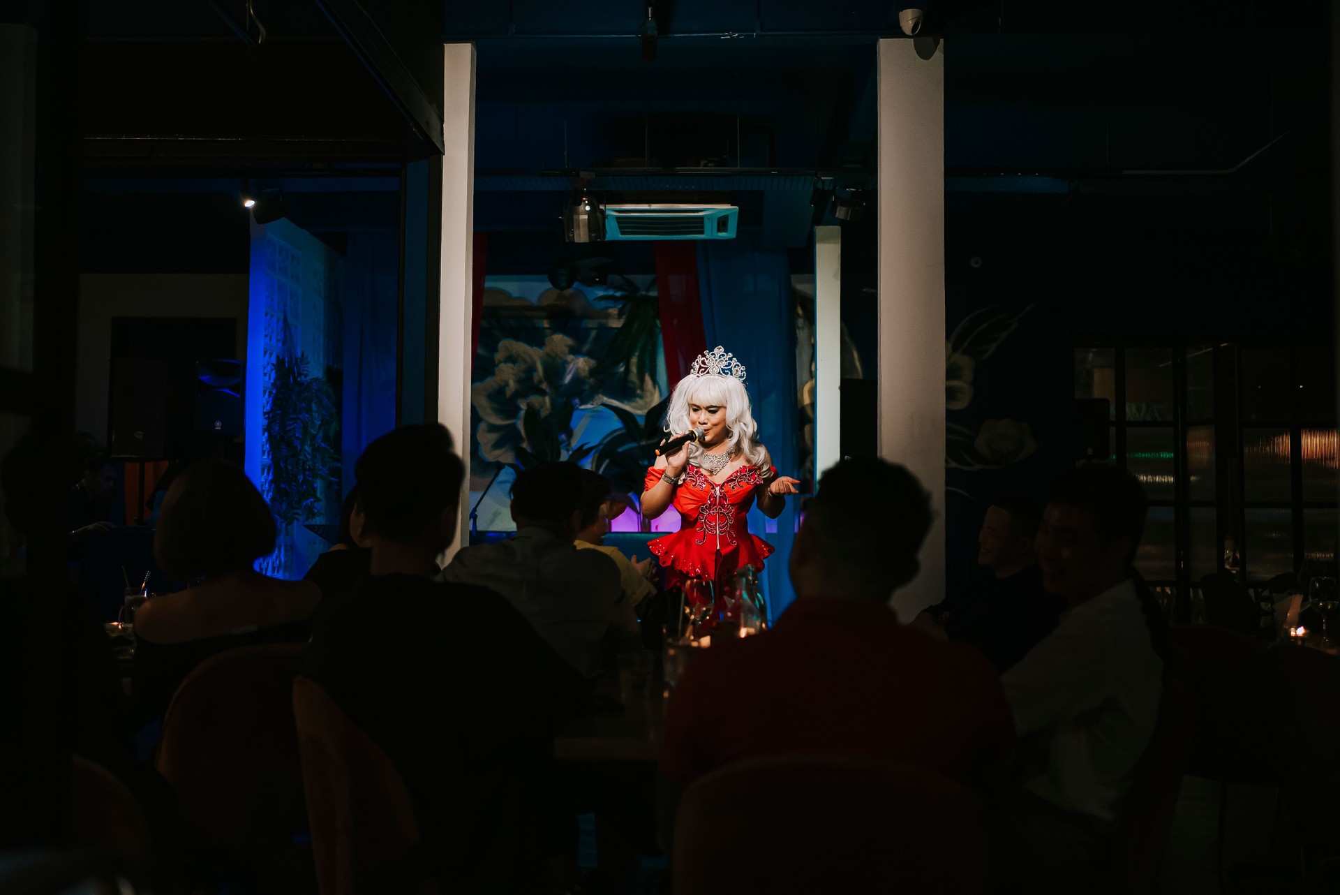 Asian drag queen in spot lit stage performance at pub with audience in silhouette